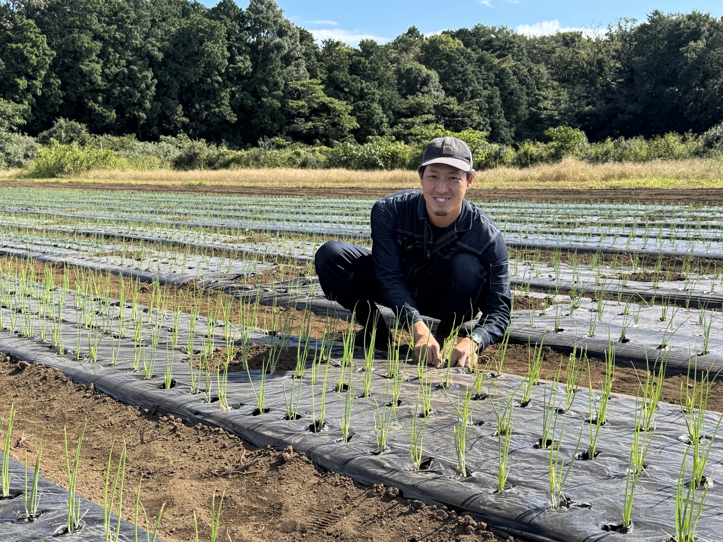 葉ネギの苗を植えた畑で、苗に手を添えてほほえむAjito Farmの代表